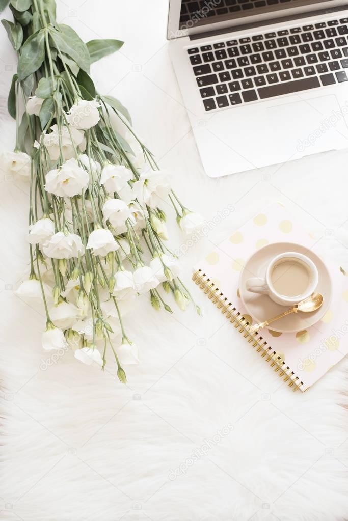 Laptop, coffee, notebook and a large bouquet white flowers on the floor on a white fur carpet. Freelance fashion comfortable femininity home workspace in flat lay style. Top view, pink and gold
