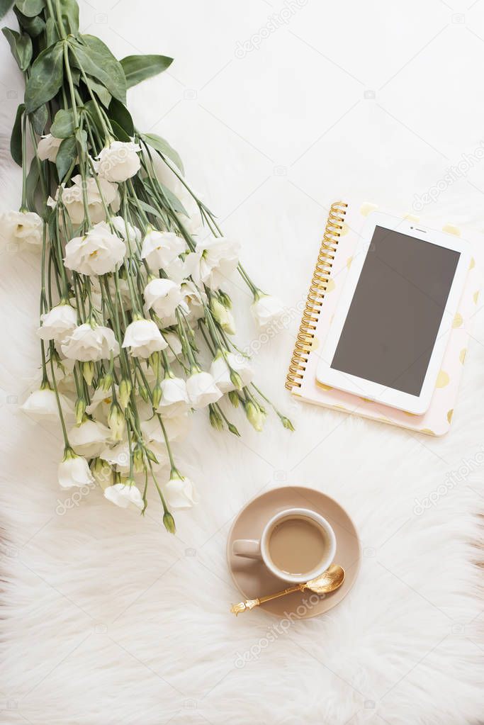 Notebook, tablet, a cup of coffee and a large bouquet white flowers on the floor on a white fur carpet. Freelance fashion comfortable femininity home workspace in flat lay style. Top view, nude and go