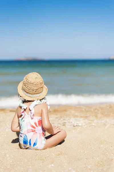 Niña pequeña con sombrero de paja y traje de baño floral mirando al mar en un día soleado. Niña sentada en la orilla del mar — Foto de Stock