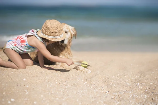 Niña, niña con sombrero de paja y traje de baño floral jugando con un perro en la playa en un día soleado. La chica y su Golden retriever sentados en la orilla del mar — Foto de Stock