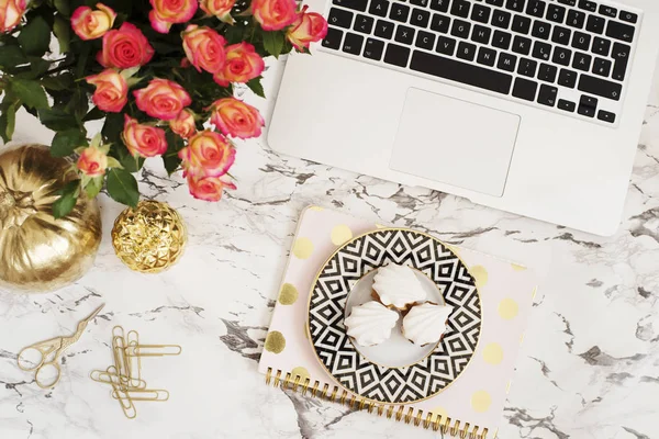 Feminine workplace concept. Freelance workspace in flat lay style with laptop, flowers, golden pineapple, notebook and paper clips on white marble background. Top view, bright, pink and gold