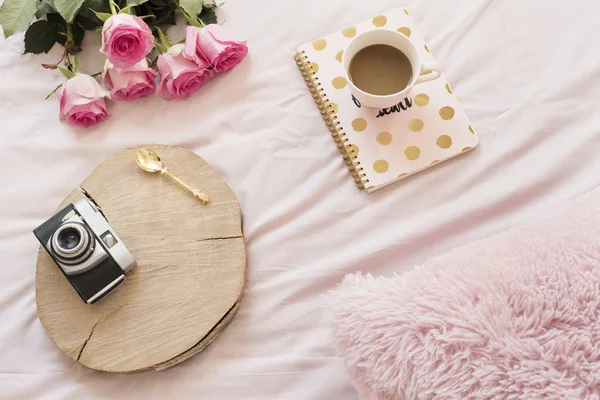 Caffè, vecchia fotocamera vintage a letto su lenzuola rosa. Rose e quaderni in giro. Freelance casa di moda femminilità spazio di lavoro in stile laico piatto — Foto Stock