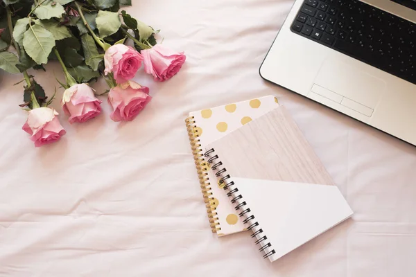 Cuadernos, portátil, ramo de rosas en la cama sobre sábanas rosadas. Espacio de trabajo de feminidad en casa de moda independiente en estilo laico plano . —  Fotos de Stock