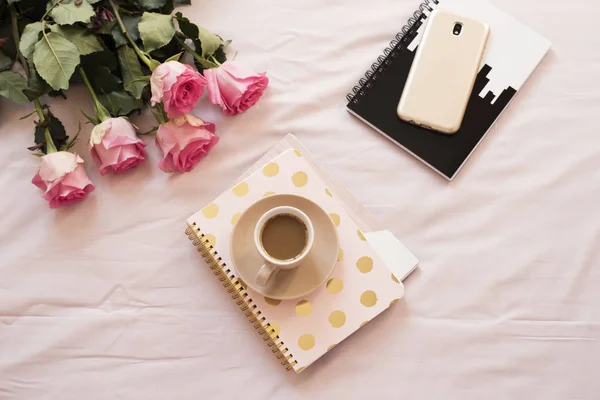 Café en la cama sobre sábanas rosas. Rosas, cuadernos, teléfonos inteligentes de oro alrededor. Espacio de trabajo de feminidad de casa de moda independiente —  Fotos de Stock