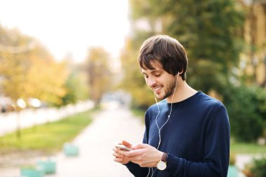 Yürüyüş ve sokak kentsel arka plan üzerinde kulaklık ile müzik dinlemek için bir akıllı telefon kullanarak mutlu genç adam