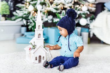 Cute little baby child boy in blue hat sitting plaing with toys at a home interior and waiting Santa on background of the modern Christmas tree clipart