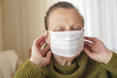 Corona virüsüne karşı korunmak için cerrahi maske takan yaşlı kadının portresi. Büyükannem oturma odasında oturuyor ve yukarı bakıyor. 