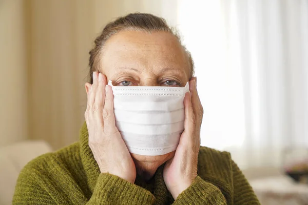 Portrait of old woman wearing surgical mask for protection against corona virus. Grandmother sitting in her living room and looking up