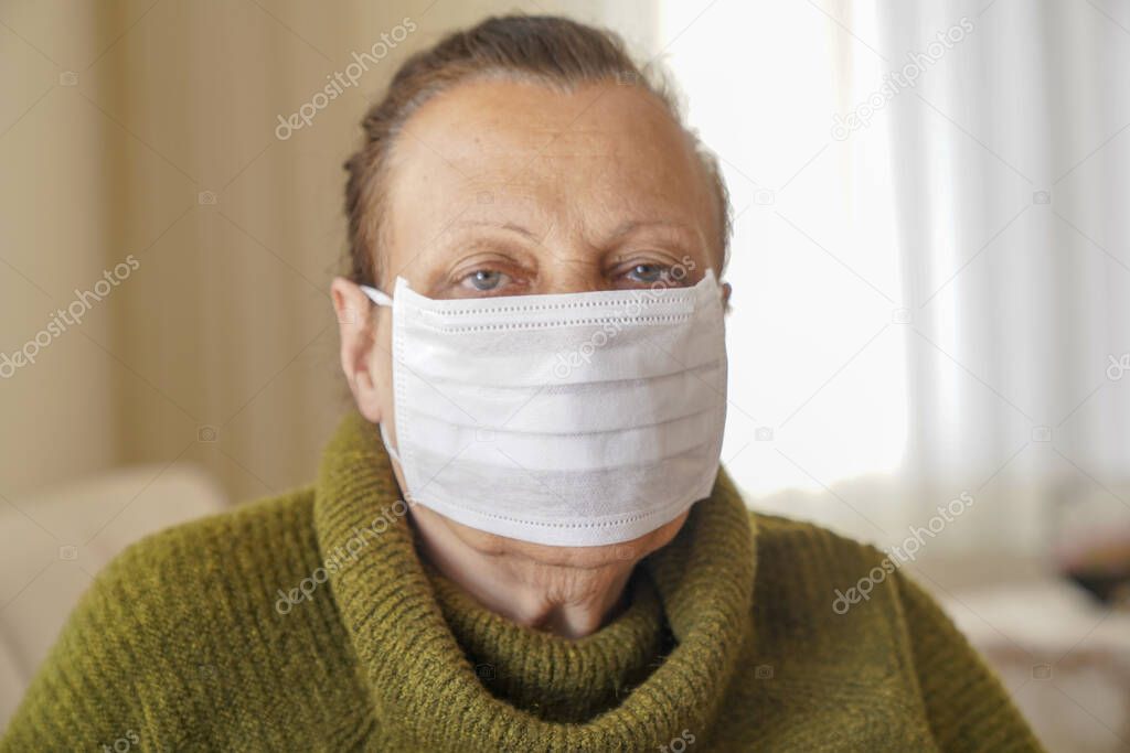 Portrait of old woman wearing surgical mask for protection against corona virus. Grandmother sitting in her living room and looking up 