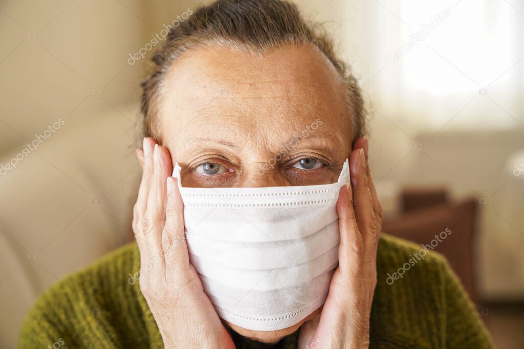 Portrait of old woman wearing surgical mask for protection against corona virus. Grandmother sitting in her living room and looking up 