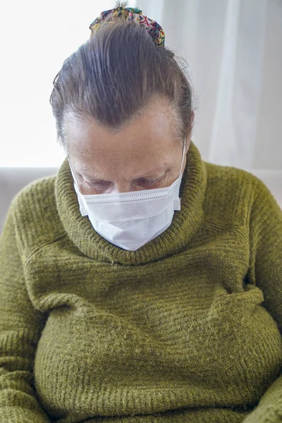 Corona virüsüne karşı korunmak için cerrahi maske takan yaşlı kadının portresi. Büyükannem oturma odasında oturuyor ve yukarı bakıyor. 