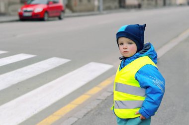 Yaya geçidinin önündeki çocuk. Küçük çocuk yaya geçidinden geçip geçemeyeceğini öğreniyor. Güvenlik nedeniyle yansıtıcı yelek giyiyor. Arkadaki araba. Çocuk konsepti. Trafik kavramı.