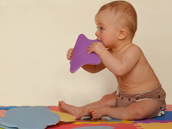 Cute baby biting foam puzzle shape is sitting on the colored mats. 