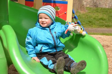 Küçük çocuk kaydırağın üstünde en sevdiği oyuncağıyla oyun alanında.