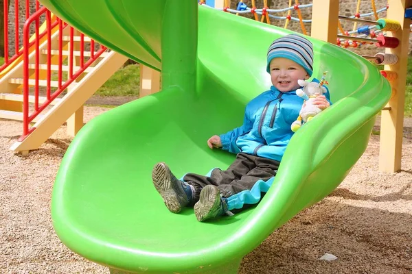 Mały chłopiec jeżdżący na zjeżdżalni z ulubioną zabawką na placu zabaw. — Zdjęcie stockowe