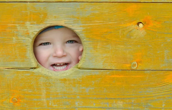 Niño feliz mirando a través de un agujero de madera en el patio de recreo. Concepto infantil . — Foto de Stock
