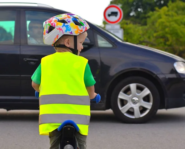 Mały chłopiec na rowerze, w odblaskowej kamizelce i kasku na drodze. Jeździ przed nim samochodem.. — Zdjęcie stockowe