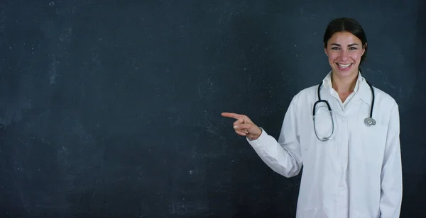 Retrato de uma bela médica (estudante) fica ao lado de uma placa preta, sobre um fundo preto. Conceito: ideias, escola, universidade, química, ciência, professores, memória, biologia, médico, faculdade . — Fotografia de Stock