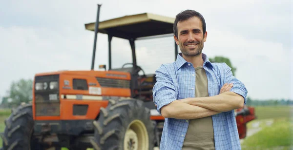 Potret seorang petani muda tampan berdiri di atas kemeja dan tersenyum di depan kamera, dengan latar belakang traktor dan alam. Konsep: ekologi biologi, lingkungan yang bersih, masyarakat yang indah dan sehat, petani . — Stok Foto
