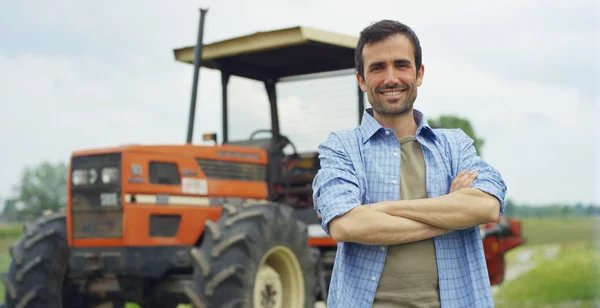 Portret van een knappe jonge boer staande in een shirt en glimlachend in de camera, op de achtergrond van een trekker en natuur. Concept: bio-ecologie, schone omgeving, mooie en gezonde mensen, boeren. — Stockfoto