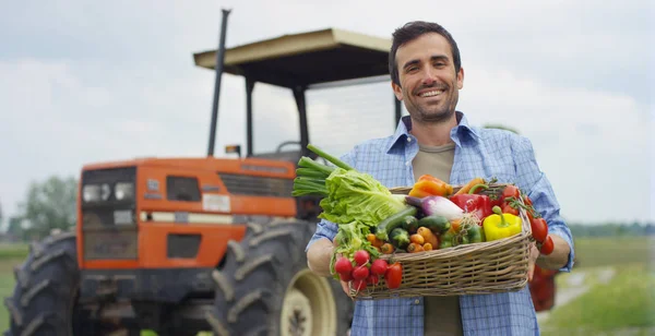 Portret szczęśliwy młody rolnik posiadający świeżych warzyw w koszu. tło z ciągnika i charakter pojęcia biologiczne, bio produktów, ekologia bio, uprawiane przez własne ręce, wegetarian, sałatki zdrowy — Zdjęcie stockowe