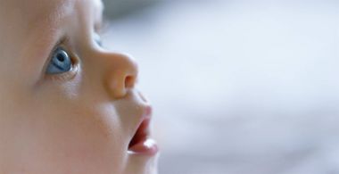 Bir bebek, büyük mavi gözleri ve açık renk saçlı, bir çocuk oturur ve bembeyaz bir battaniye var gülümsüyor, beyaz zemin üzerine annesi bakar.