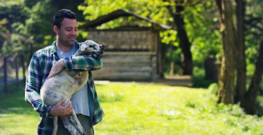 Bir kuzu genç yavrusu tutan bir genç yakışıklı ve gülümseyerek çiftçi, takip etmek ve kavram doğa ve bir ahır arka plan üzerinde canlı hayvan bakmakta deneyime sahiptir: ekoloji, çiftlik hayvanları, bio, tarım