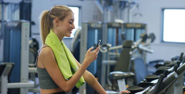 Joven chica hermosa en un gimnasio, sacude las piernas en un simulador de ciclismo, con un teléfono en la mano. El concepto: amar los deportes, asistir a un gimnasio, una nutrición adecuada, un cuerpo delgado, estar sano . — Foto de Stock