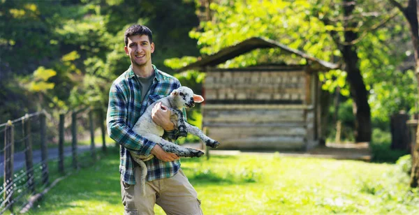 Un jeune fermier beau et souriant, tenant un jeune ourson d'agneau, a l'expérience de suivre et de prendre soin des animaux vivants, sur le fond de la nature et d'une grange, concept : écologie, bétail, bio, agriculture — Photo