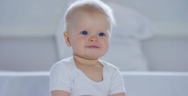 A baby, a boy with large blue eyes and light-colored hair, sits and smiles on a snow-white blanket, looks at her mother, on a white background. clipart