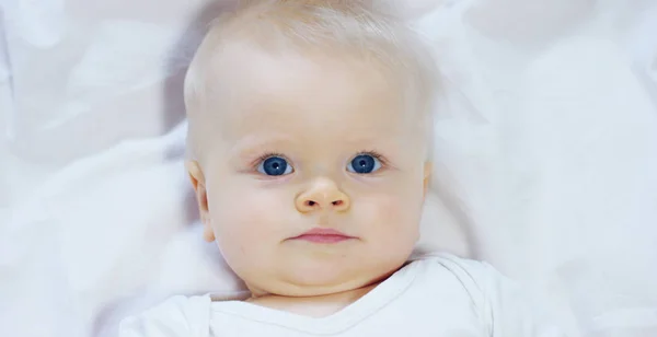 Seorang bayi, seorang anak laki-laki dengan mata biru besar dan rambut berwarna terang, duduk dan tersenyum di atas selimut putih-salju, menatap ibunya, dengan latar belakang putih . — Stok Foto
