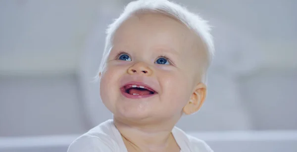 Ребенок, мальчик с большими голубыми глазами и светлыми волосами, сидит и улыбается на белоснежном одеяле, смотрит на мать, на белом фоне . — стоковое фото