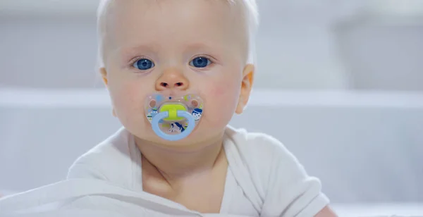 A baby, a boy with large blue eyes and light-colored hair, sits and smiles on a snow-white blanket, looks at her mother, on a white background. — Stock Photo, Image