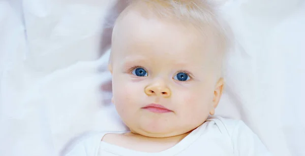 Seorang bayi, seorang anak laki-laki dengan mata biru besar dan rambut berwarna terang, duduk dan tersenyum di atas selimut putih-salju, menatap ibunya, dengan latar belakang putih . — Stok Foto