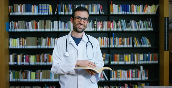 Seorang dokter wanita muda dan cantik di perpustakaan tersenyum bahagia dan memegang buku setelah melakukan pencarian dan setelah belajar. Konsep: pendidikan, potret, perpustakaan, dan perawatan medis dan kesejahteraan — Stok Foto