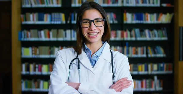 A young and beautiful female doctor in a library smiling happy and holding books after doing a search and after studying. Concept: educational, portrait, library, and medical care and welfare — Stock Photo, Image