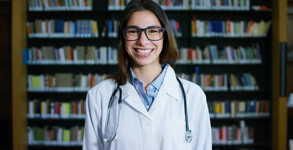 Μια νεαρή και όμορφη γυναίκα γιατρό σε μια βιβλιοθήκη χαμογελώντας ευτυχισμένη και κρατώντας βιβλία μετά από να κάνει μια αναζήτηση και μετά τις σπουδές του. Έννοια: εκπαιδευτικό, πορτραίτο, βιβλιοθήκη, και ιατρική περίθαλψη και πρόνοια — Φωτογραφία Αρχείου