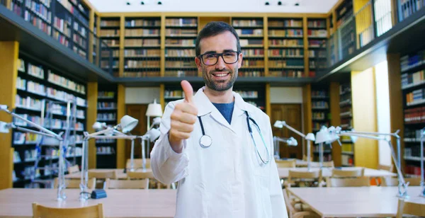 Une jeune et belle femme médecin dans une bibliothèque souriante heureuse et tenant des livres après avoir fait une recherche et après avoir étudié. Concept : éducation, portrait, bibliothèque, soins médicaux et bien-être — Photo