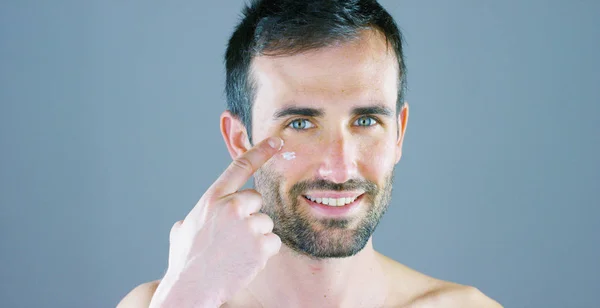 Un hombre muy guapo sonríe y pone la crema facial y el contorno de ojos anti-arrugas y anti-envejecimiento para el cuidado de su cuerpo y conoce la belleza. Concepto: belleza, crema, belleza — Foto de Stock