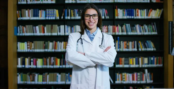 Una joven y hermosa doctora en una biblioteca sonriendo feliz y sosteniendo libros después de hacer una búsqueda y después de estudiar. Concepto: educación, retrato, biblioteca, atención médica y bienestar —  Fotos de Stock