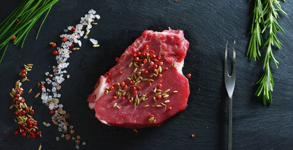 Steak daging segar yang lezat di atas meja dengan garam, rosemary, bawang putih, dan tomat di latar belakang hitam, pemandangan atas. Konsep: produk segar & alami, produk bio, produk daging, organik, nutrisi . — Stok Foto
