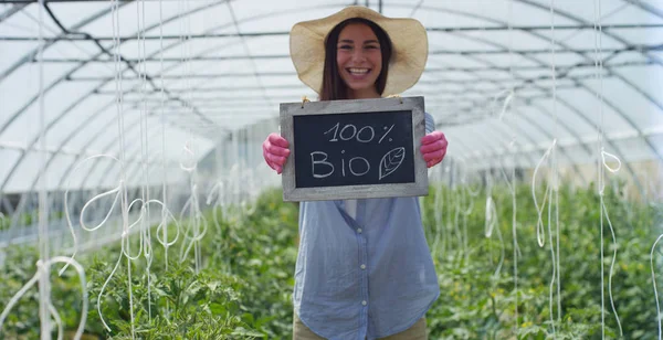 Uma menina bonita em um chapéu de palha e usando luvas de borracha rosa, segura uma placa preta, em uma estufa. Conceito: produtos biológicos, produtos naturais, frescos, deliciosos, frutas, legumes, crescer, água, plantas — Fotografia de Stock