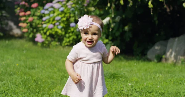 Bayi perempuan yang baru lahir mencoba untuk membuat langkah-langkah pertama di sebuah taman pada hari yang cerah mengenakan gaun bayi penari merah muda dengan bunga di kepalanya ekspresi bagus seperti bayi lucu konsep sekolah tari menyenangkan — Stok Foto