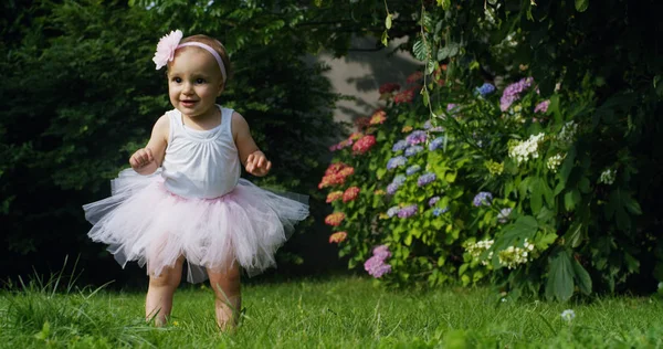 Bayi perempuan yang baru lahir mencoba untuk membuat langkah-langkah pertama di sebuah taman pada hari yang cerah mengenakan gaun bayi penari merah muda dengan bunga di kepalanya ekspresi bagus seperti bayi lucu konsep sekolah tari menyenangkan — Stok Foto