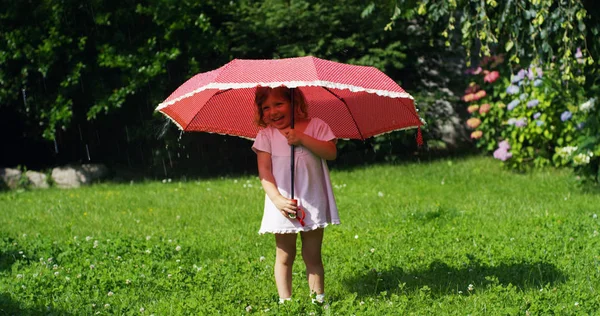 Menina no jardim brincando com os pais se escondendo sob um guarda-chuva vermelho contra espirrar água e olhando para a câmera. conceito de segurança e poupança de energia — Fotografia de Stock