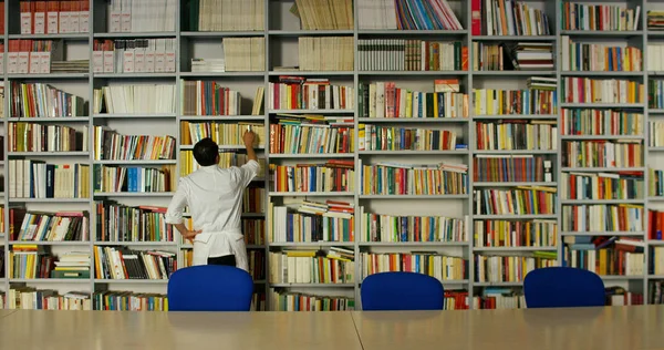 Un jeune et beau médecin dans une bibliothèque souriant heureux et tenant des livres après avoir fait une recherche et après avoir étudié. Concept : éducation, portrait, bibliothèque, soins médicaux et bien-être — Photo