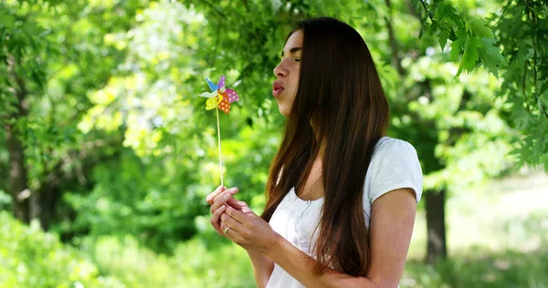 En un día de sol y viento, una hermosa chica feliz en la naturaleza verde sopla en un molinete colorido ayudado por la energía del viento fresco. concepto de felicidad y energía eólica renovable.Resolución 6k — Foto de Stock