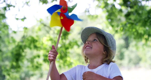 Em um dia do sol e do vento, uma menina bonita pequena feliz na natureza verde sopra em um pinwheel colorido ajudado pela energia do vento fresco. conceito de felicidade e energia eólica renovável.6k resolução — Fotografia de Stock