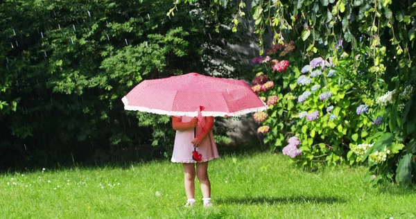 Niña en el jardín jugando con los padres escondidos bajo un paraguas rojo contra salpicaduras de agua y mirando a la cámara. concepto de seguridad y ahorro de energía — Foto de Stock