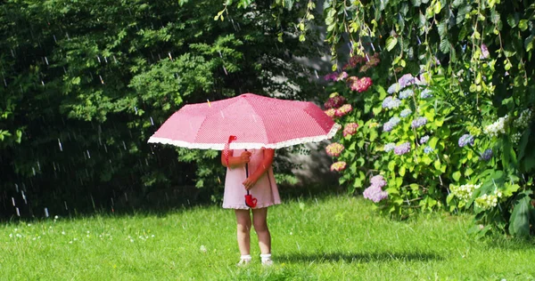 Kleines Mädchen im Garten, das mit Eltern spielt, die sich unter einem roten Regenschirm vor dem plätschernden Wasser verstecken und in die Kamera schauen. Konzept der Sicherheit und Energieeinsparung — Stockfoto